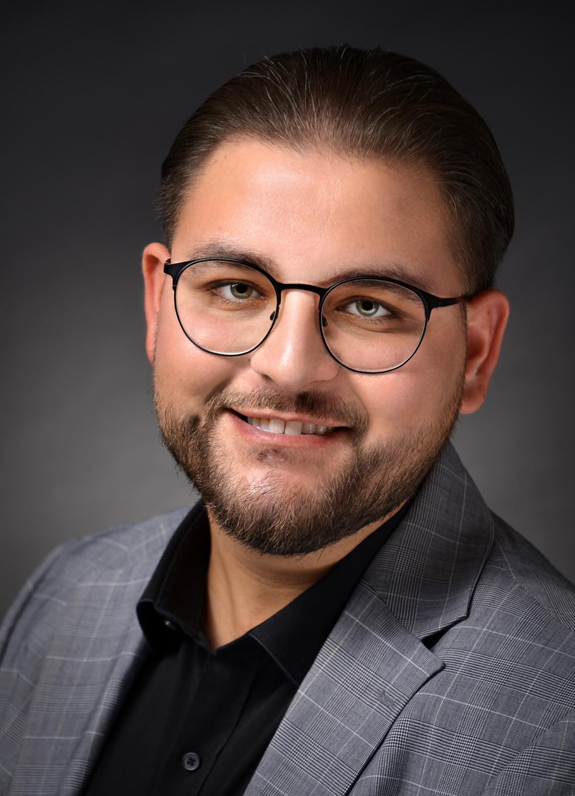 Männlich gelesene Person mit Brille, lächelnd in die Kamera blickend